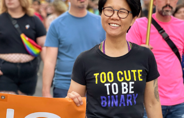 A person in a black shirt with colourful text smiles as they hold an orange and white banner. They are marching with others on the road.