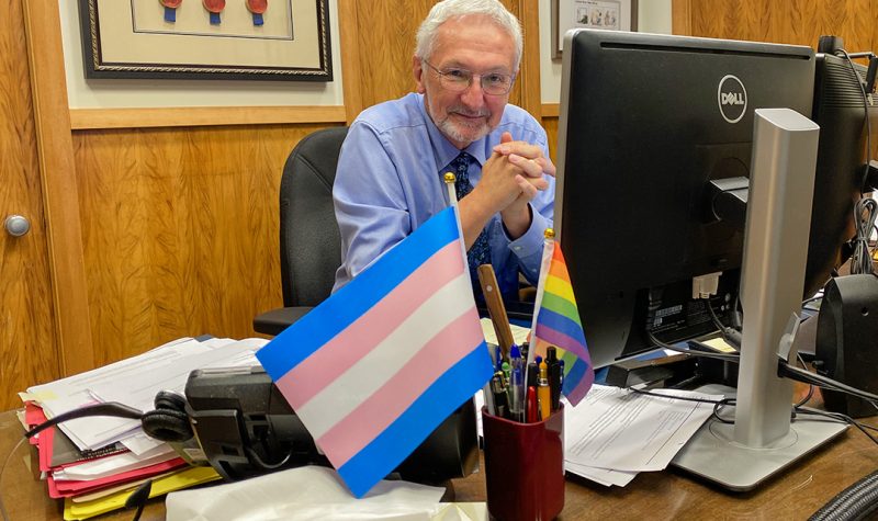 A man with white hair and a beard sits in an office with the trans and rainbow Pride flag in a mug on his desk.