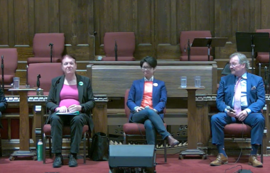 People sitting in a row on a stage with a rainbow flag on the left and an ice sculpture of a person's bust on the right.