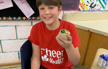 Child holding a wrap in a classroom. There is a red and white checkered sandwich paper in their lap.