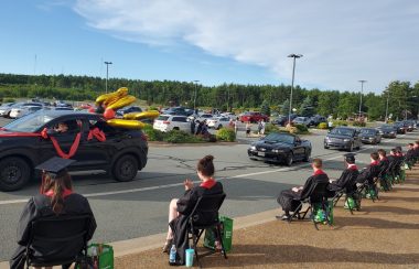 A grad drive-thru parade is seen with a number of vehicles driving by grads sitting in chairs spaced apart on a sunny day