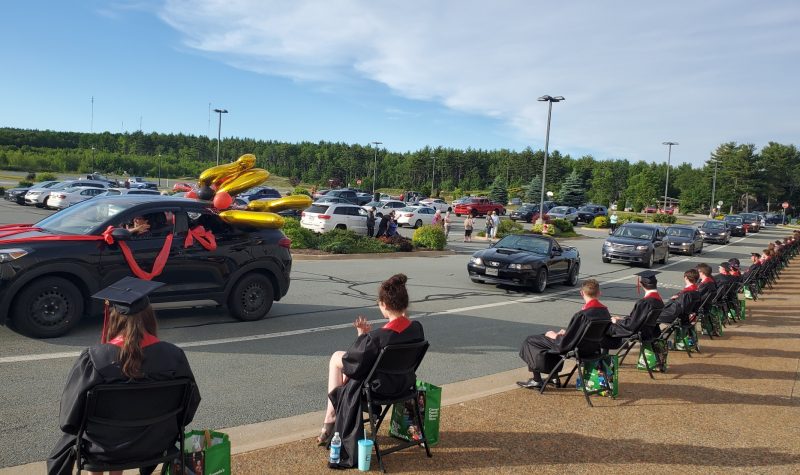 A grad drive-thru parade is seen with a number of vehicles driving by grads sitting in chairs spaced apart on a sunny day
