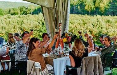 Pictured are some people enjoying a previous Grande Tablée event. They are seated at a round table covered with a white table cloth and wine glasses. They are all holding their wine glasses up in the air to do a toast.