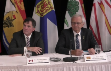Two men sit at a table with microphones and various flags stand behind them including New Brunswick, Nova Scotia, PEI and Newfoundland.