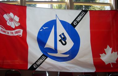 A flag with a blue sailboat, two horseshoes, a diagonal line with the words “49th parallel”, a dogwood flower, and a maple leaf on a red and white Canadian flag-style background. 