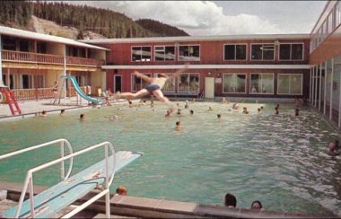 A pool is busy with people swimming during the day at a hot springs resort in northern BC. A young person is jumping off of the diving board preparing to do an epic belly flop.