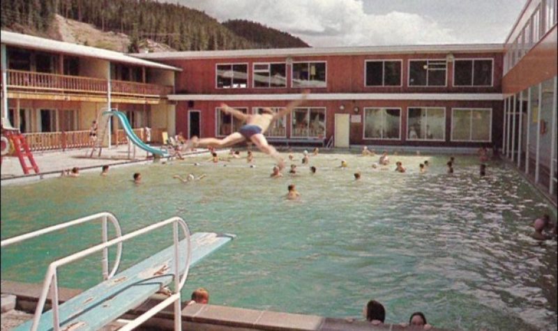 A pool is busy with people swimming during the day at a hot springs resort in northern BC. A young person is jumping off of the diving board preparing to do an epic belly flop.