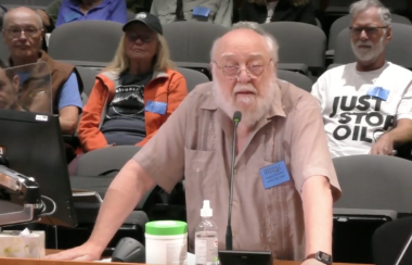 A bald white man with white hair and a white beard wearing a tan shirt and a blue nametak stands in front of a microphone with people sitting behind him includng one wearing a shirt with Just Stop Oil printed on it