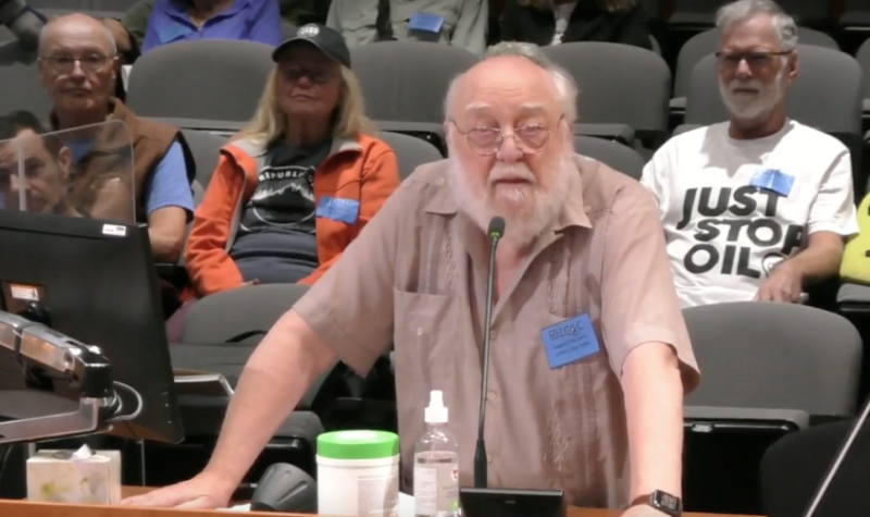 A bald white man with white hair and a white beard wearing a tan shirt and a blue nametak stands in front of a microphone with people sitting behind him includng one wearing a shirt with Just Stop Oil printed on it