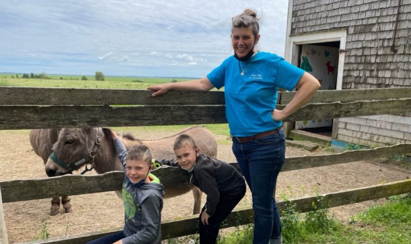 Laura Hunter leans on a fence with her two grandchildren. Behind the fence are two donkeys.