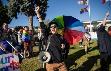 A person holding a rainbow umbrella over their shoulder and a megaphone over the other smiling with their fist in the air