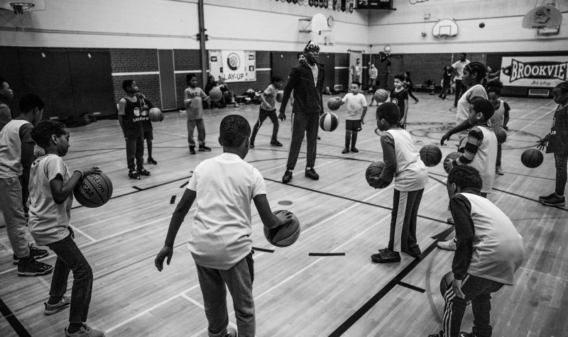 Photo of Lay-Up pre-pandemic, with students dribbling basketballs around their coach in a school gym.