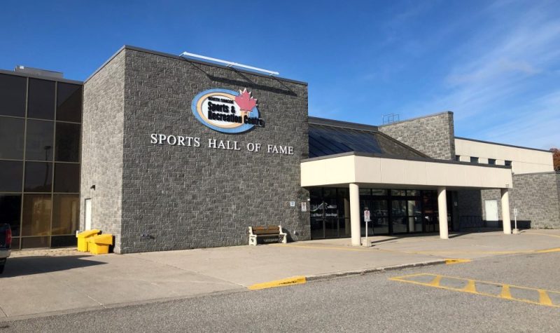 On peut voir la façade du North Simcoe Sport and Recreation Center de Midland. C'est un bâtiment moderne fait de brique et de béton gris. L'affiche du Centre est visible sur la façade