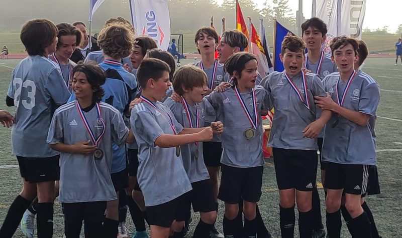 Equipe d'une quinzaine de jeunes joyeux en chandail gris portant leurs medailles des jeux de l'acadie.