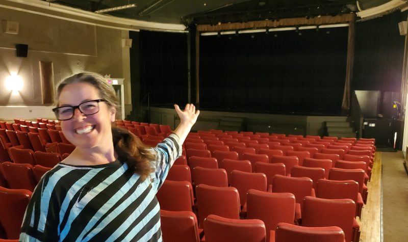 A woman stands at the back of a theatre gesturing towards the stage from the back rows of seats.