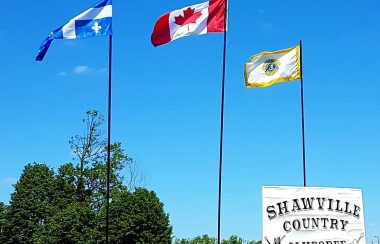 A photo of the entrance to the Shawville Lions Country Jamboree grounds with a large white sign and the Quebec, Canadian and Lions Club flags in the background.