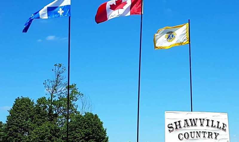 A photo of the entrance to the Shawville Lions Country Jamboree grounds with a large white sign and the Quebec, Canadian and Lions Club flags in the background.