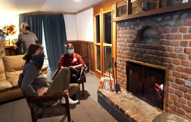 Two people sit in front of a lighted brick fireplace with one person standing behind, all wearing masks in a dimly lit room