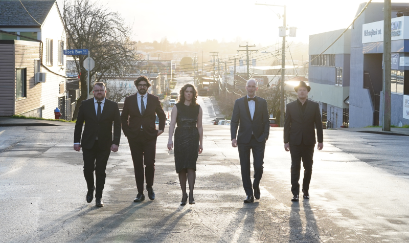Five bandmates dressed formally, walk down the street with the sun beaming against their backs.