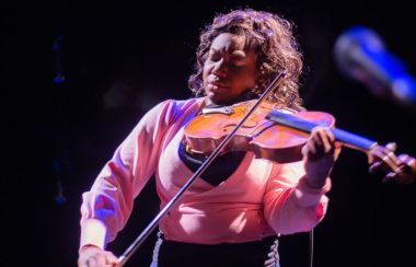 A Black woman with curly, shoulder-length hair and wearing a pink blouse plays a violin with her eyes closed.