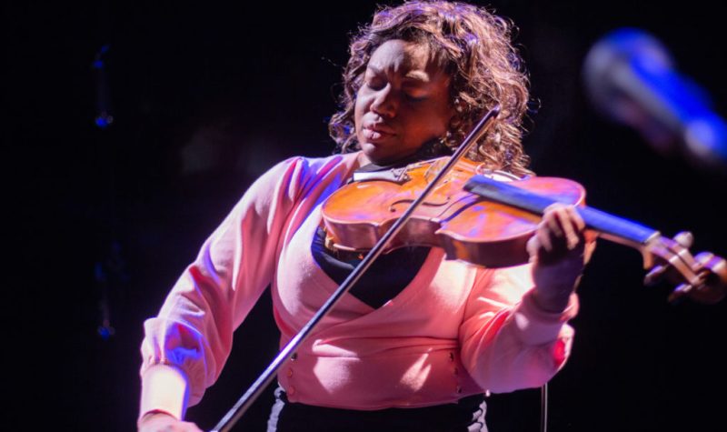 A Black woman with curly, shoulder-length hair and wearing a pink blouse plays a violin with her eyes closed.