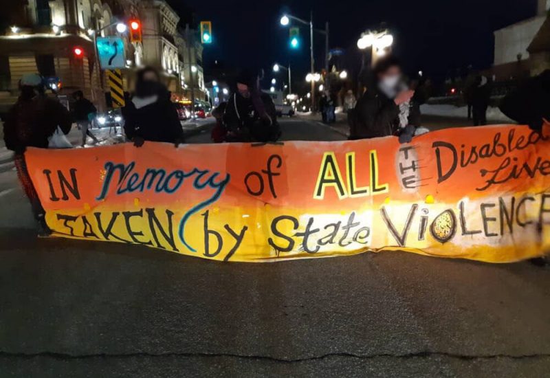 An image of an orange and yellow banner held up by five protestors. The banner says, 