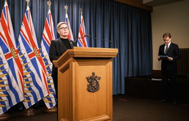 Mary Ellen Turpel-Lafond stands at a wooden podium at a press conference with a row of BC flags behind her.