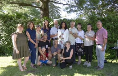 A group of people stand for a photo outside on a sunny day. Some are holding glasses of wine with greenery serving as the background.