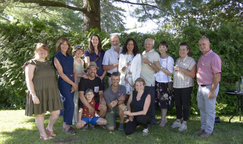 A group of people stand for a photo outside on a sunny day. Some are holding glasses of wine with greenery serving as the background.