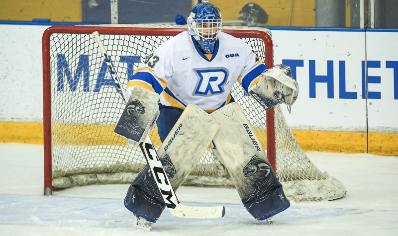 A person with blue, white and yellow goalie equipment on on a hockey rink indoors.