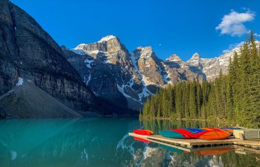 A picture of Moraine Lake, with the sun lighting up the crystal blue glacier water. The iconic mountain peaks shine in the background. Weather is clear, with a little snow on the mountains