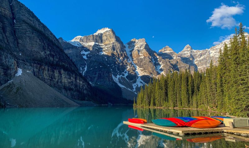 A picture of Moraine Lake, with the sun lighting up the crystal blue glacier water. The iconic mountain peaks shine in the background. Weather is clear, with a little snow on the mountains