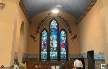 The altar area of a former church. A large stained glass window shines in the background, which is surrounded by stylized writing. Before it are artifacts and items belonging to war front and home front efforts of past wars.