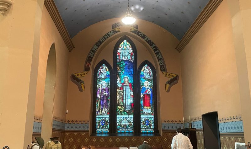 The altar area of a former church. A large stained glass window shines in the background, which is surrounded by stylized writing. Before it are artifacts and items belonging to war front and home front efforts of past wars.