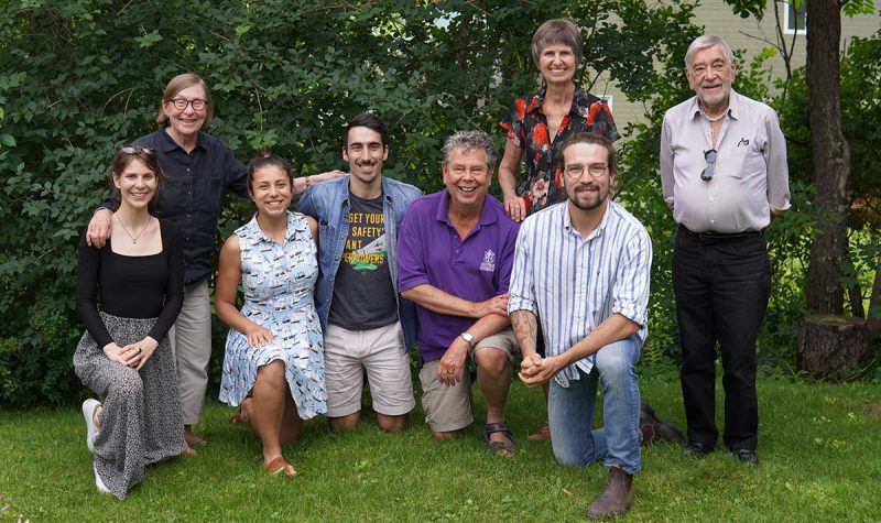 A group of 8 people kneel on the grass against a backdrop of trees.