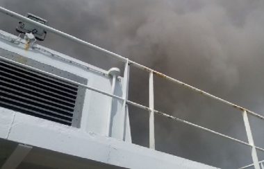 Dark smoke fills the sky above the white railing and the white upper portions of a ship, as seen from below, apparently from a lower deck.