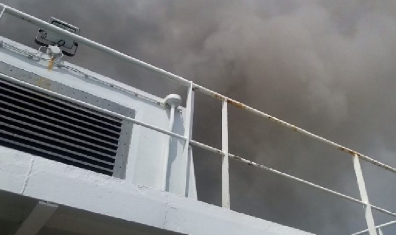 Dark smoke fills the sky above the white railing and the white upper portions of a ship, as seen from below, apparently from a lower deck.
