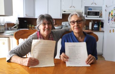 Deux femmes assises à une table de cuisine sourient à la caméra en tenant des feuilles dactylographiées.