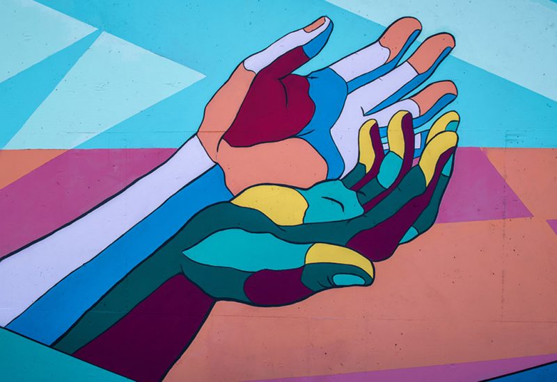 An image of a painting depicting outstretched hands. They are cupped and painted in blue, orange red, yellow, green and brown.