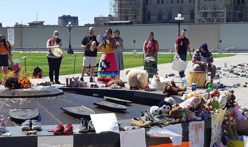 An image of Jackie Shigwadjah in a ribbon skirt holding a bullhorn. Behind her is a line of drummers and in front of here is a temporary memorial to the Kamloops 215.