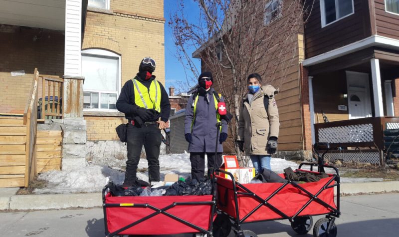 Three people wearing masks, two wearing high visibility vests, standing behind two red wagons. These wagons are filled with supplies for the homeless.