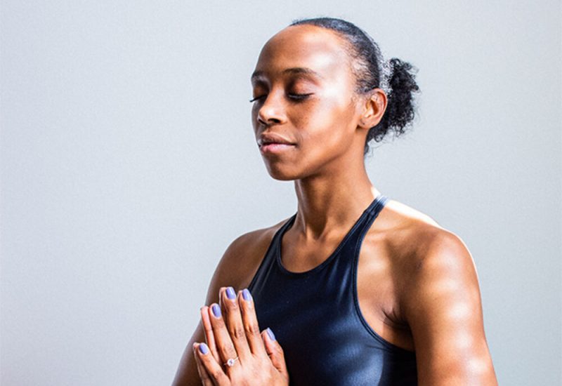 A Black woman meditates with her eyes closed and her hands pressed together at heart centre against a grey background