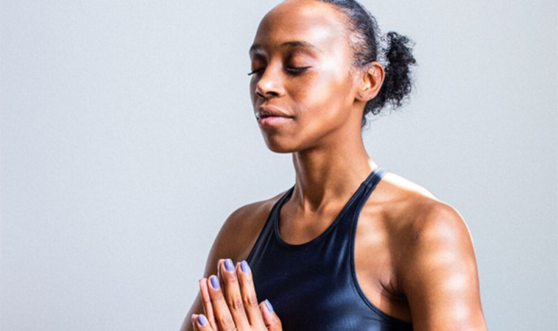 A Black woman meditates with her eyes closed and her hands pressed together at heart centre against a grey background