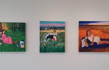 Three bright and vivid art pieces of Lana Whiskeyjack exhibit, first picture is a couple sisters taking a break from berry picking sitting against a tree and second a woman in front of a ceremonial grounds, last picture is an Indigenous lady laying on her side with a orange field with an oxen in background