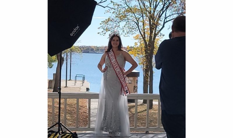Young indigenous teenage girl wearing a white formal gown and a crown with a sash that reads, Miss Teen Canada Petite 2021-2022