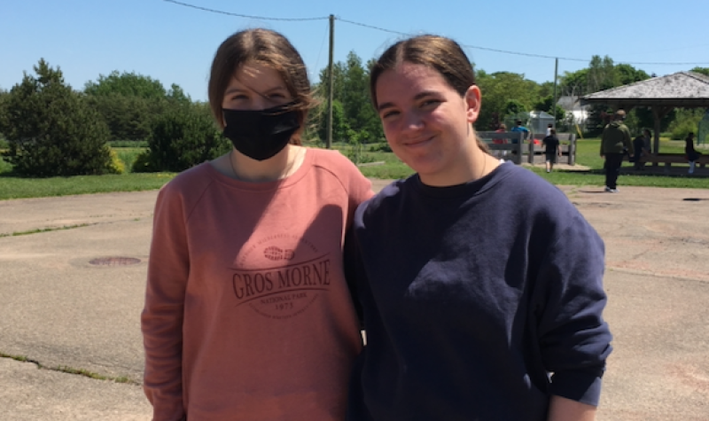 Two young women standing outside in a school yard, one wearing a face mask.
