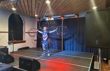 On aperçoit l'artiste autochtone Makhena Rankin-Guerin performant la « danse des cerceaux ». Elle se tient debout sur la scène intérieure de la Salle Roland-Desroches en tenant plus de 15 cerceaux avec tout son corps.