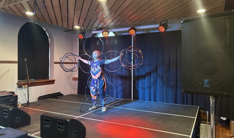 On aperçoit l'artiste autochtone Makhena Rankin-Guerin performant la « danse des cerceaux ». Elle se tient debout sur la scène intérieure de la Salle Roland-Desroches en tenant plus de 15 cerceaux avec tout son corps.