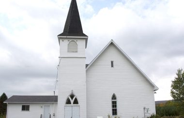 Pictured is a photograph taken of the outside of the Potton Valley Church in Mansoville. The church is white and is set against the cloudy sky.