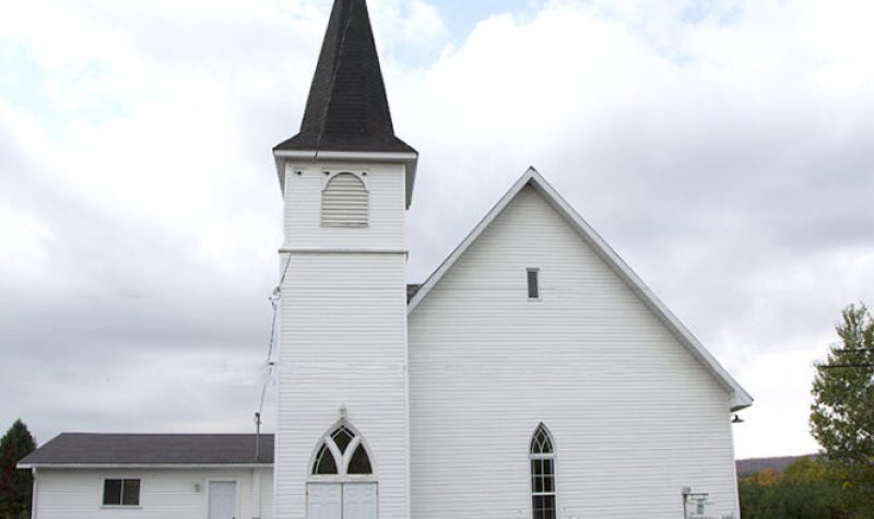 Pictured is a photograph taken of the outside of the Potton Valley Church in Mansoville. The church is white and is set against the cloudy sky.